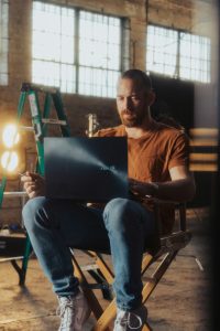 Ein Mann sitzt konzentriert auf einem Regiestuhl mit Laptop auf dem Schoss in einem kreativen Arbeitsumfeld mit Studiolicht und Leiter im Hintergrund. Flexible Arbeitsmodelle wie diese bieten Menschen mit ADHS in der Schweiz die Möglichkeit, ihren Beruf individuell zu gestalten.