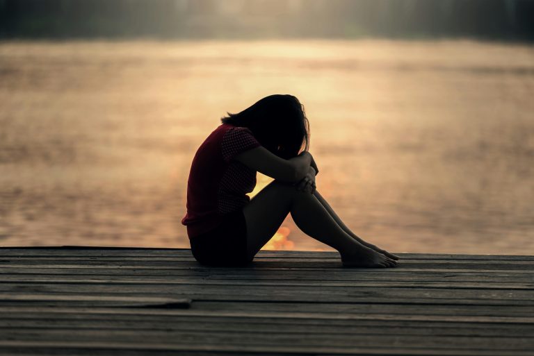 Eine junge Frau sitzt alleine am Steg eines Sees und hat den Kopf auf die Knie gelegt. Die untergehende Sonne spiegelt sich im Wasser, was eine melancholische Stimmung erzeugt. Dieses Bild symbolisiert emotionale Überforderung – ein häufiges Begleitsymptom bei ADHS in der Schweiz, das oft übersehen wird.