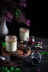 Zwei Gläser mit Overnight-Oats und kleine Gläser mit Beeren stehen auf einem rustikalen Holztisch, umgeben von Blumen und Schokoladenstücken. Besonders bei ADHS Schweiz kann ein nährstoffreiches Frühstück den Start in den Tag verbessern. Die Kombination aus gesunden Kohlenhydraten, Eiweiss und Antioxidantien fördert die Konzentration.