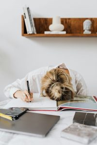 Eine junge Frau hat den Kopf auf ein offenes Buch gelegt, während sie noch einen Stift in der Hand hält – sie wirkt erschöpft und überfordert. Der Arbeitsbereich ist chaotisch, was auf eine Überlastung hindeutet. In der ADHS Schweiz Diagnostik sind solche Symptome typische Anzeichen für kognitive Überforderung.