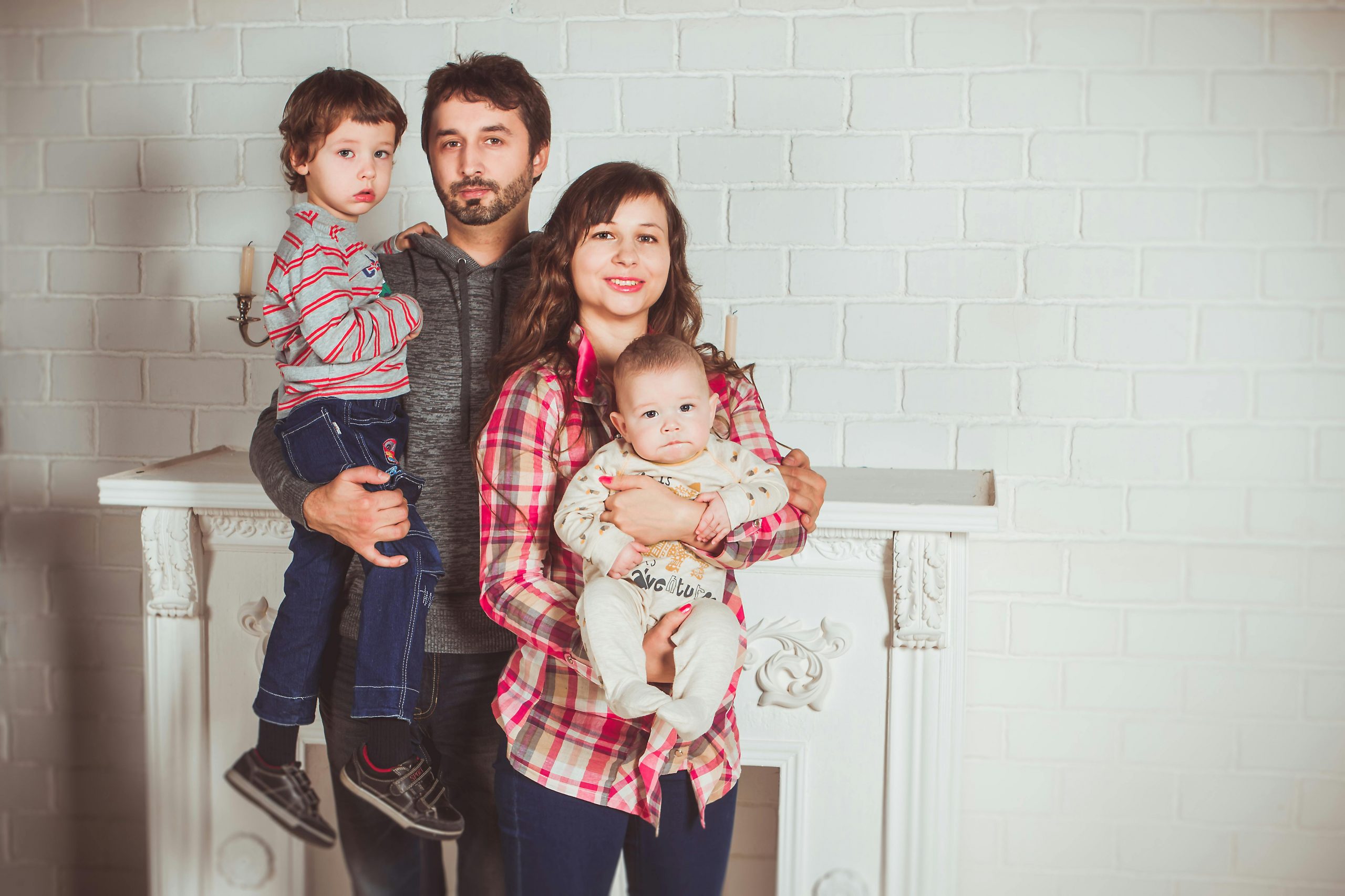 Eine junge Familie posiert liebevoll vor einer weißen Backsteinwand – die Eltern tragen ihre beiden Kinder auf dem Arm. Ein harmonisches Familienleben ist auch mit ADHS im Beruf in der Schweiz möglich – mit den richtigen Strukturen und Unterstützungssystemen.