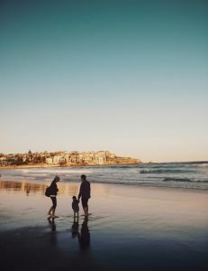 Eine Familie spaziert barfuß am Strand entlang, während im Hintergrund eine malerische Küstenstadt in der Abendsonne leuchtet. Solche gemeinsamen Momente helfen dabei, Beruf und Familie besser zu vereinbaren – besonders wenn ADHS im Beruf in der Schweiz eine Rolle spielt.