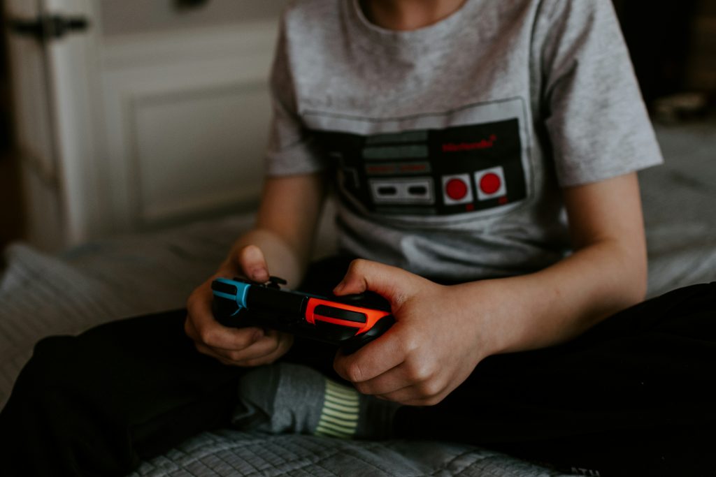 Ein Kind sitzt auf dem Bett und spielt mit einem bunten Nintendo-Controller, bekleidet mit einem Retro-Gaming-Shirt. Der Fokus liegt auf dem Controller in den kleinen Händen. Im Kontext von ADHS Schweiz ist die Bildschirmzeit bei Kindern ein zentrales Thema – insbesondere in Hinblick auf Aufmerksamkeit und Impulskontrolle.