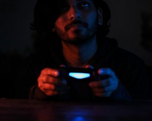 Ein junger Mann hält einen leuchtenden Controller und schaut nachdenklich ins Dunkel – das blaue Licht wirft eine fast meditative Stimmung. Es entsteht ein Kontrast zwischen innerer Unruhe und äußerer Stille. Für Betroffene von ADHS Schweiz kann das Zocken sowohl Rückzug als auch Herausforderung bedeuten – je nach Nutzung.
