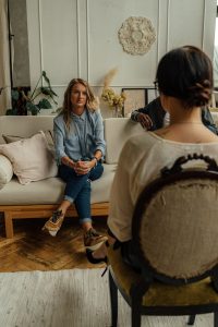 Eine junge Frau sitzt mit verschränkten Händen auf einem Sofa, gegenüber eine Person mit Notizblock. Die ruhige Umgebung lässt Raum für Reflexion und Vertrauen. Auch in der Schweiz ist die ADHS-Gruppentherapie oft in kleine Gruppen oder Paargespräche integriert, um individuelle Bedürfnisse besser zu berücksichtigen.