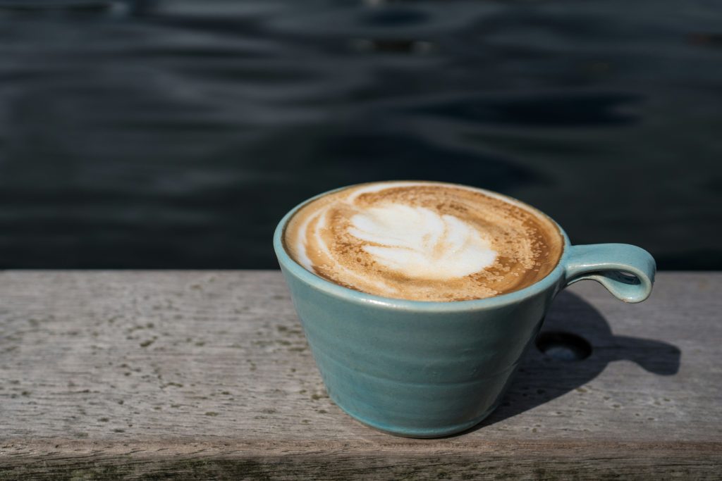 Eine Tasse Cappuccino mit schöner Latte-Art steht auf einer Holzbank am Wasser. Das ruhige Ambiente symbolisiert eine entspannte Pause im Alltag. Auch Menschen mit ADHS Schweiz können von bewussten Ruhepausen profitieren.