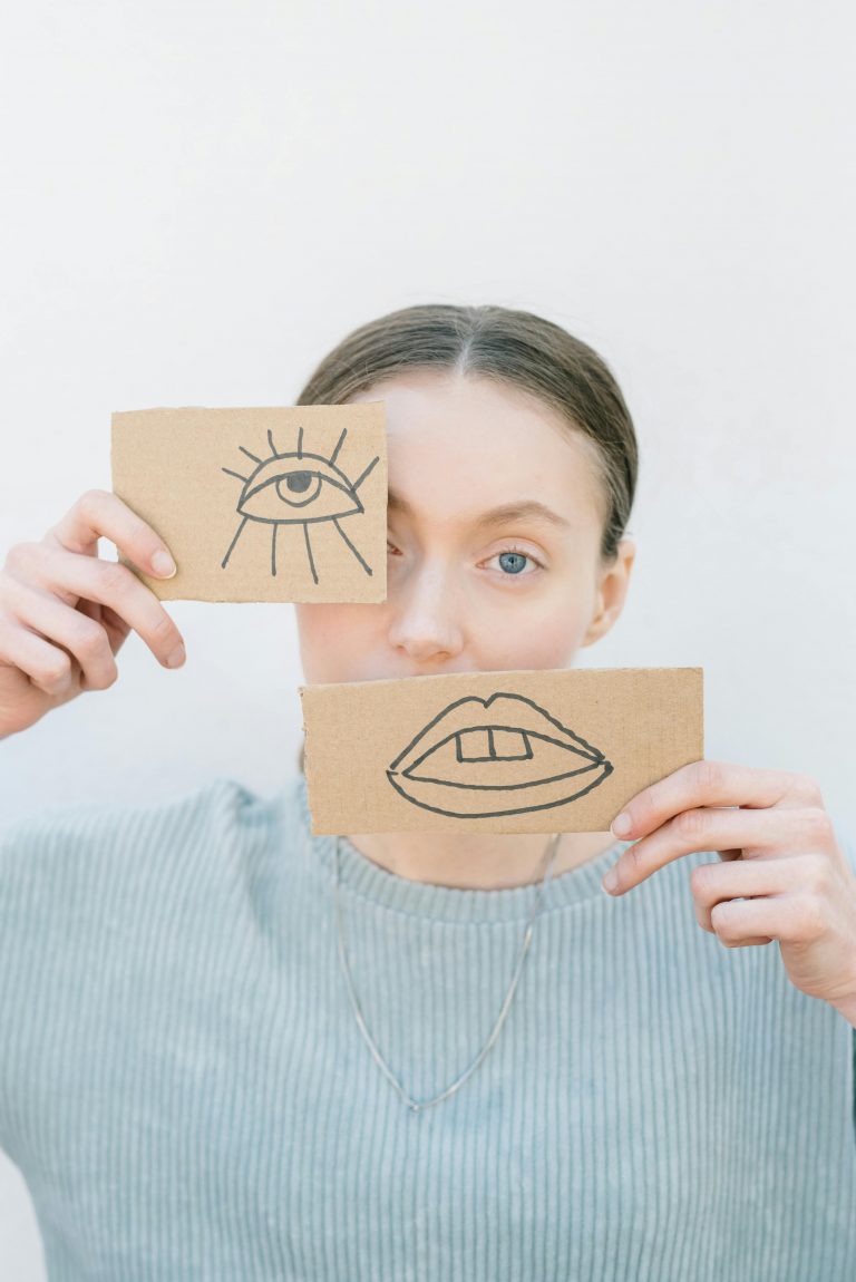 Eine junge Frau hält Pappstücke mit einem gezeichneten Auge und Mund vor ihr Gesicht – ein Symbol für die Schwierigkeit, Emotionen bei ADHS oder Autismus auszudrücken.