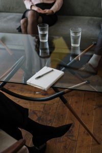 Ein Tisch mit zwei Gläsern, einem Notizbuch und einer Brille steht im Vordergrund, während sich zwei Personen im Hintergrund unterhalten. Der Fokus liegt auf Details, die Vertrauen und Professionalität symbolisieren. Ein typisches Setting einer ADHS Therapie in der Schweiz.