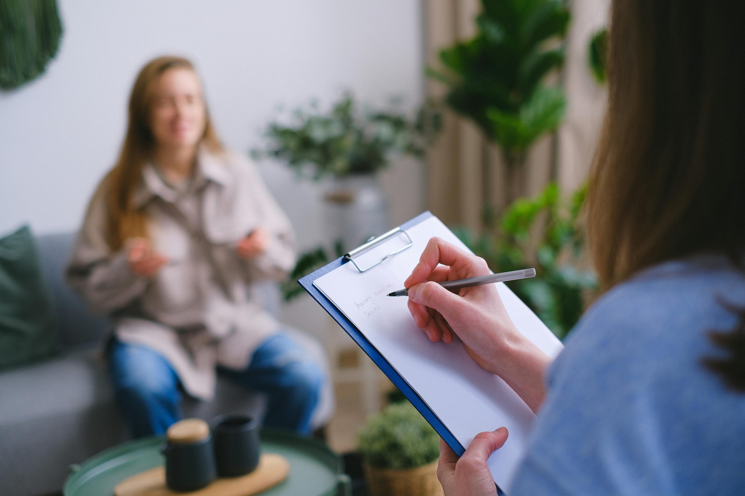 Eine Therapeutin notiert auf einem Klemmbrett, während im Hintergrund eine Frau gestikulierend spricht. Die Umgebung ist freundlich und grün gestaltet. Die Szene repräsentiert eine moderne und empathische ADHS Therapie in der Schweiz.