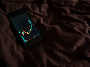 Ein Smartphone liegt auf einem braunen Bettlaken und zeigt eine Börsen-App mit Kerzenchart. Die nächtliche Nutzung elektronischer Geräte kann bei ADHS Schweiz den Schlafrhythmus erheblich stören. Schlafhygiene ist ein wichtiger Bestandteil der ADHS-Therapie.