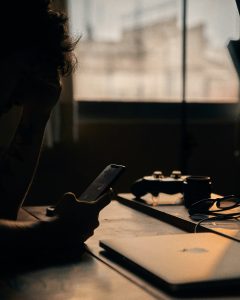 Eine Person sitzt im Dunkeln an einem Schreibtisch und starrt auf ihr Smartphone. Auf dem Tisch liegen ein Laptop, eine Spielkonsole und eine Brille – Zeichen von Reizüberflutung und digitaler Ablenkung. Dieses Bild steht sinnbildlich für die Herausforderungen bei ADHS in der Schweiz im Umgang mit Bildschirmzeit und Konzentration.