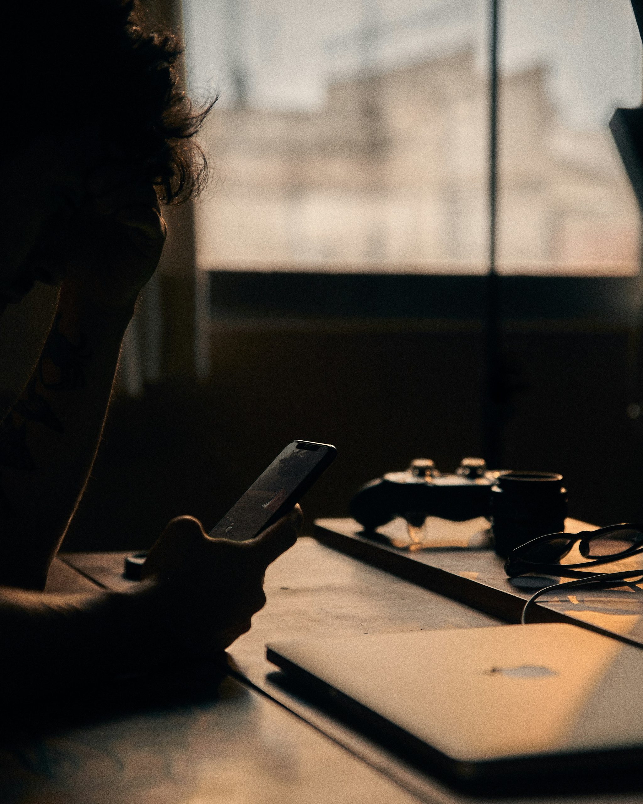 Eine Person sitzt im Dunkeln an einem Schreibtisch und starrt auf ihr Smartphone. Auf dem Tisch liegen ein Laptop, eine Spielkonsole und eine Brille – Zeichen von Reizüberflutung und digitaler Ablenkung. Dieses Bild steht sinnbildlich für die Herausforderungen bei ADHS in der Schweiz im Umgang mit Bildschirmzeit und Konzentration.