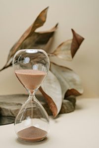 Eine elegante Sanduhr mit feinem, braunem Sand steht auf einem hellen Untergrund, im Hintergrund sind getrocknete Blätter zu sehen. Im Kontext von ADHS Schweiz wird oft thematisiert, wie bewusste Zeitstrukturierung helfen kann, Stress und Überforderung zu vermeiden. Visuelle Timer wie Sanduhren sind einfache, aber effektive Hilfsmittel.