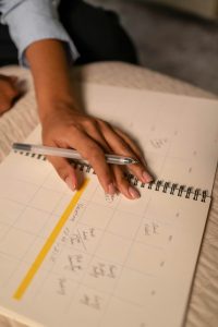 ine Hand mit Kugelschreiber ruht auf einem Kalender mit handschriftlichen Einträgen und einer gelb markierten Woche. Strukturierte Zeitplanung ist für Personen mit ADHS im Beruf in der Schweiz ein wichtiger Schlüssel zur besseren Selbstorganisation.