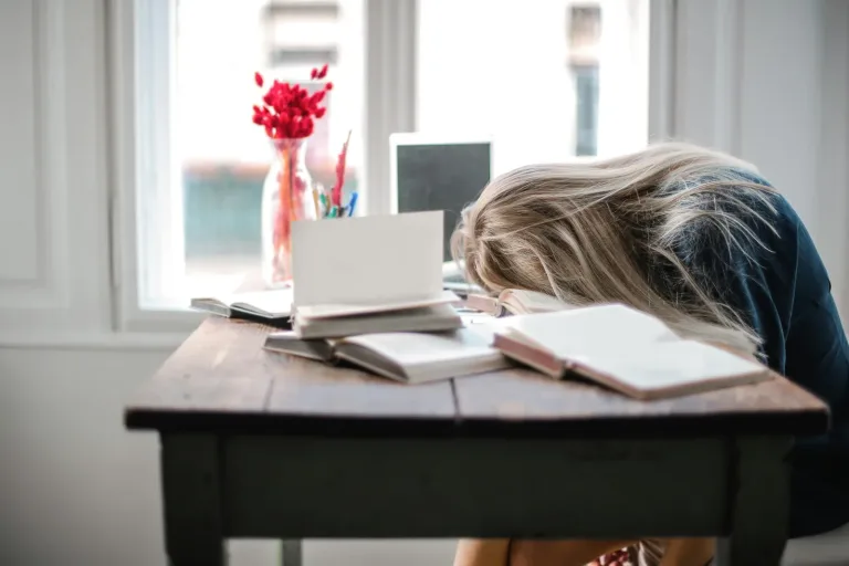 Frau liegt erschöpft mit dem Kopf auf dem Schreibtisch neben Büchern und Notizbuch