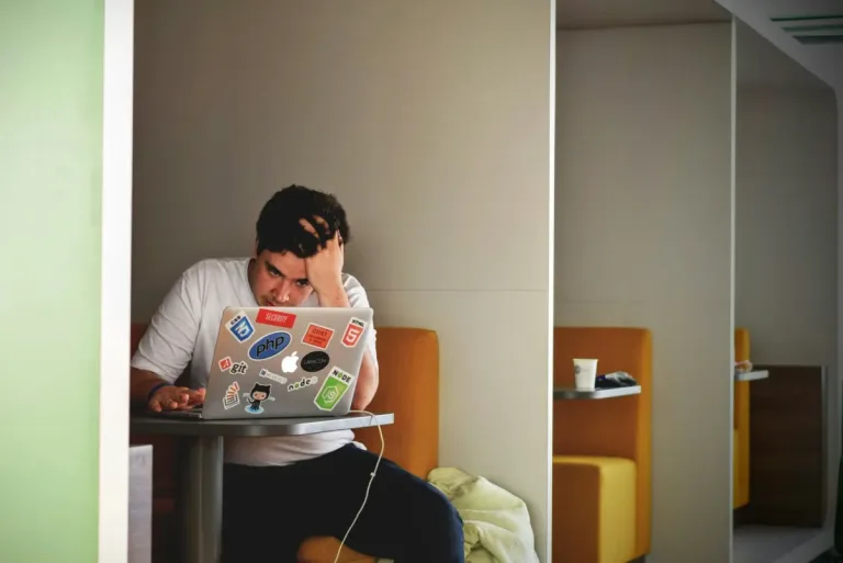 Ein junger Mann sitzt mit gesenktem Kopf vor einem Laptop, der über und über mit Entwickler-Stickern beklebt ist. Er wirkt frustriert und überfordert – ein Gefühl, das viele Erwachsene mit ADHS Schweiz im Berufsleben kennen. Das Bild verdeutlicht den Bedarf nach passenden Tools und gezielter Entlastung.