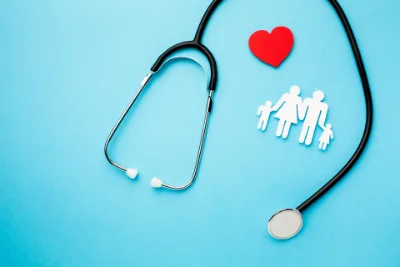 Ein Stethoskop liegt auf einem blauen Untergrund, z.B. einem Tisch. Stilistisch danaeben eine Familie und darüber ein Herz: Absicherung über die Krankenversicherung.