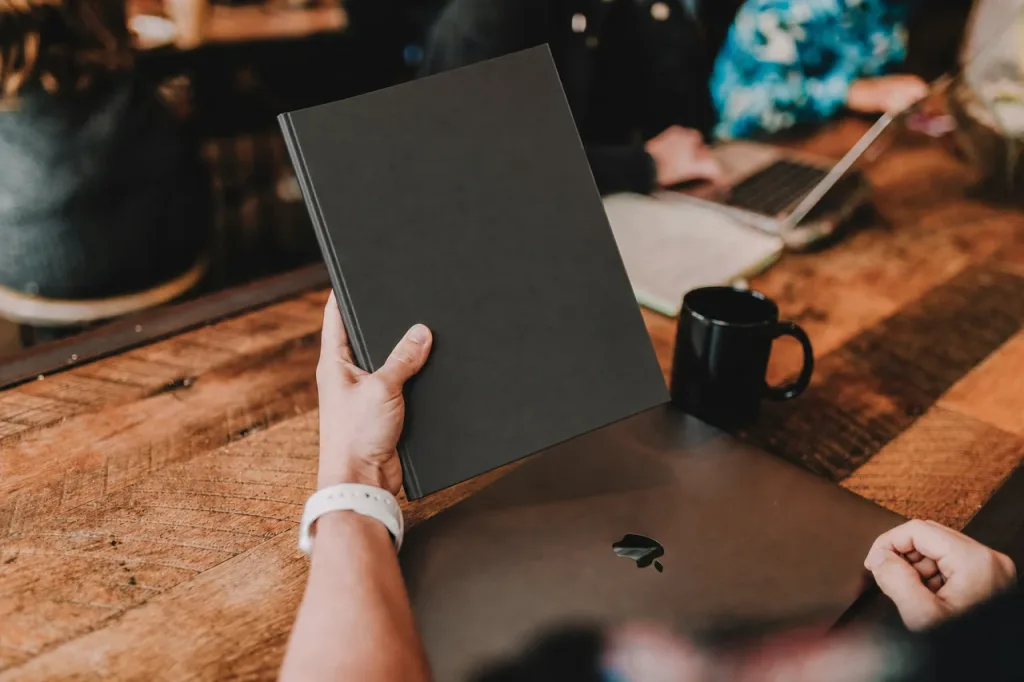 Person hält ein Notizbuch über einem Laptop an einem Holztisch