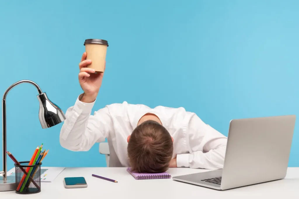 Ein Mann in weissem Hemd liegt erschöpft auf seinem Schreibtisch, während er einen Pappbecher in die Höhe hält. Neben ihm stehen ein Laptop, ein Handy und Büromaterialien. Müdigkeit und Konzentrationsprobleme sind typische Herausforderungen für Menschen mit ADHS Schweiz.